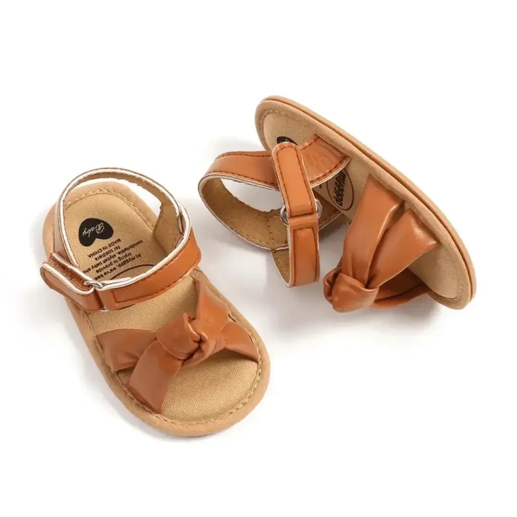 Par de sandálias infantis marrom com laço frontal, tira ajustável no tornozelo e solado bege, estilo confortável para bebê menina."