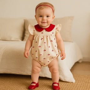 Bebê de aproximadamente 7 meses em pé, sorridente, usando body bege com estampa de cerejinhas, gola vermelha e sapatinhos vermelhos, em cenário aconchegante.