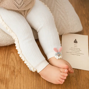 Bebê usando meia-calça infantil canelada branca com babado lateral e aplique de coelhinho rosa e cinza, em cenário aconchegante de madeira.