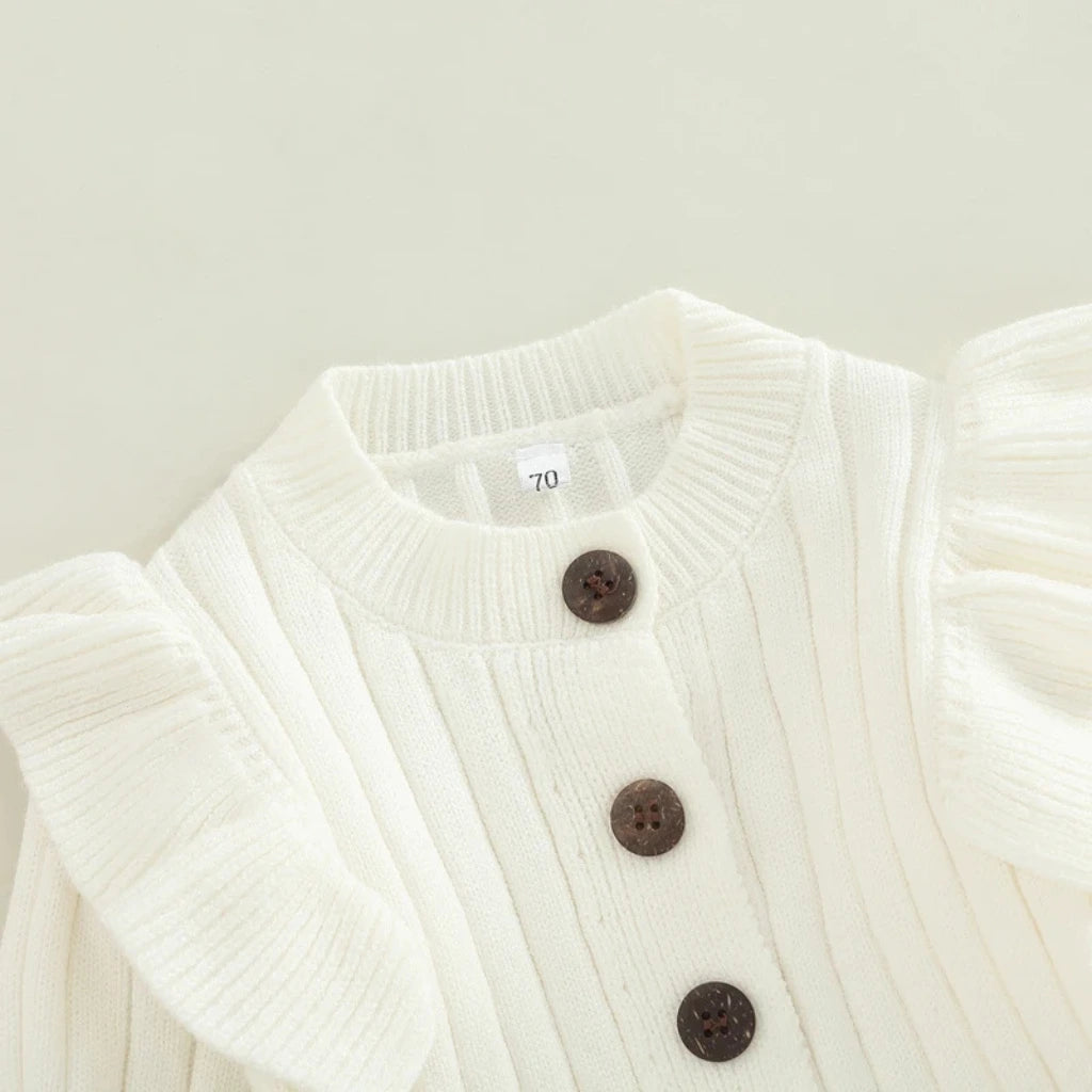 Detalhe do macacão bebê menina branco de tricô, mostrando botões frontais escuros e babados delicados nos ombros.
