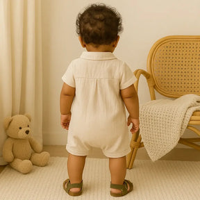 Vista traseira de bebê de 7 meses com pele morena, vestindo macacão bege de manga curta e sandálias verdes, em pé em ambiente aconchegante com ursinho e cadeira de vime ao fundo.