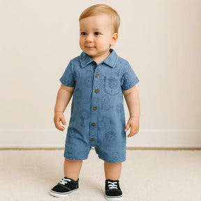 Bebê menino loiro usando macacão jeans azul com estampa de leões e sapatos pretos, em pé sobre tapete claro em ambiente aconchegante.