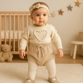 Bebê de aproximadamente 6 meses em pé, usando romper tricot bege com coração em relevo, faixa de cabelo combinando, meia calça branca e sapatinhos, em cenário aconchegante.