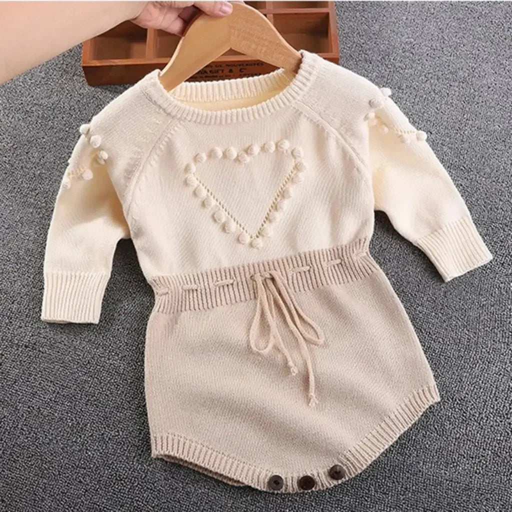Romper tricot bebê menina bege com detalhe de coração em relevo, manga longa, laço na cintura e botões funcionais