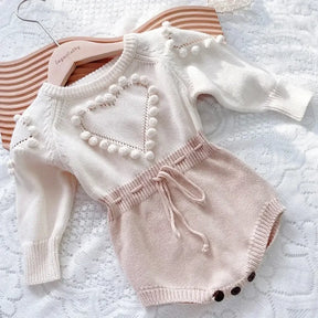 Romper tricot bebê menina bege com detalhe de coração em relevo, manga longa, laço na cintura e botões funcionais