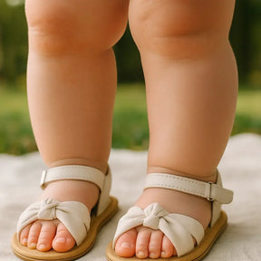 Close-up dos pés de um bebê usando sandálias infantis pretas com laço, solado bege e tira ajustável no tornozelo, em um cenário ao ar livre sobre tecido claro.