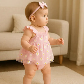 Bebê menina em pé, usando body rosa com saia de tule estampada de margaridas e faixa de cabelo com laço rosa, em cenário aconchegante de sala